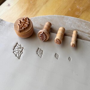 May include: Four light-colored wooden stamps with cylindrical handles, each featuring a different sized oak leaf design at the base. The stamps are arranged next to their corresponding leaf-shaped impressions pressed into a flat slab of light gray clay.