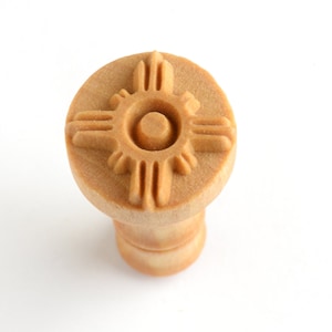 May include: A wooden dog chew toy shaped like a cylinder with a raised design of a sun symbol on the top. The chew toy is light brown in color.