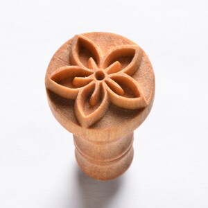 May include: A wooden stamp with a carved flower design. The stamp is brown and has a round top with a handle.