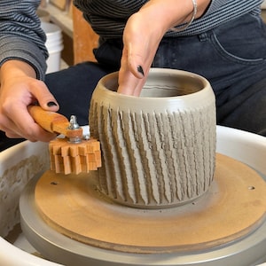 May include: A cylindrical gray clay vessel centered on a pottery wheel, featuring a textured exterior pattern created by a wooden rolling tool with a gear-like wheel held against the side to impress vertical, repeating grooves into the soft clay.
