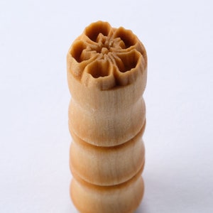 May include: A light brown, wooden stamp with a floral design on top. The stamp is cylindrical, with a stacked appearance. The top features a detailed flower pattern, while the body is smooth and rounded. The stamp is likely used for decorative purposes.