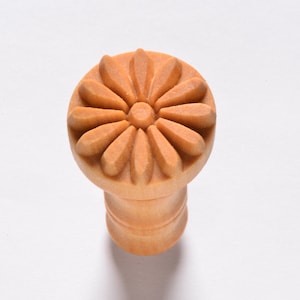 May include: A wooden stamp with a daisy design. The stamp is light brown and has a raised design.