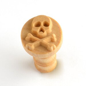 May include: A wooden stamp featuring a skull and crossbones design. The stamp has a round head with the skull and bones carved into the wood. The handle is cylindrical and made of the same light-colored wood.