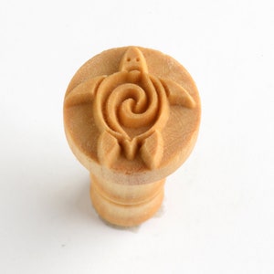 May include: A wooden stamp with a raised design of a turtle with a spiral shell. The stamp is light brown in color and has a round top with a cylindrical base.