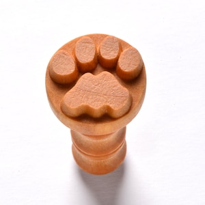 May include: A wooden stamp with a paw print design. The stamp is light brown and features a detailed paw print on the top, with a cylindrical handle. The stamp is likely used for crafts or adding a paw print to various surfaces.