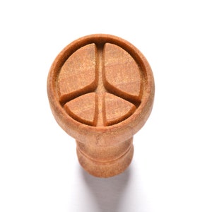 May include: A wooden peace sign shaped bottle stopper. The stopper is brown and has a smooth finish.