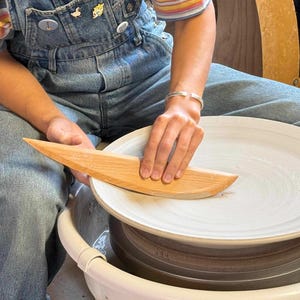 May include: A light-colored wooden pottery rib tool held against a flat, circular, unglazed clay plate centered on a white pottery wheel. The plate features concentric circular grooves from the shaping process, and the tool is used for smoothing.