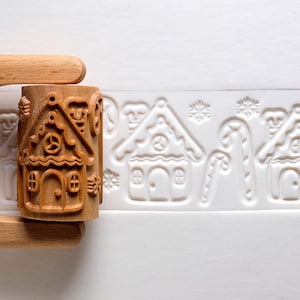 May include: A light brown wooden rolling pin with a cylindrical barrel featuring carved patterns of gingerbread houses, candy canes, and snowflakes. The rolling pin is shown pressing into a white dough surface, leaving a clear embossed impression.
