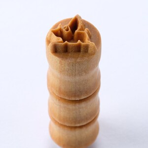 Könnte beinhalten: Ein Stempel aus Holz mit einem geschnitzten Design eines stilisierten Blattes. Der Stempel ist aus hellbraunem Holz gefertigt und hat eine zylindrische Form. Er ist in drei Abschnitte unterteilt, die jeweils einen leicht unterschiedlichen Braunton aufweisen.