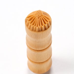 Puede incluir: Un sello de madera con un diseño de flor tallada. El sello es cilíndrico y tiene un color marrón claro.