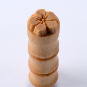 May include: A wooden stamp with a carved flower design. The stamp is light brown and has a cylindrical shape.
