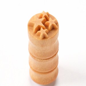 May include: A wooden stamp with three star shapes carved into the top. The stamp is made of light brown wood and is stacked on top of two other wooden pieces.