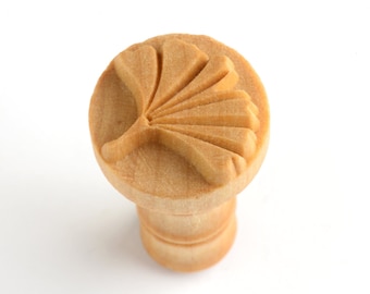 May include: A wooden stamp with a carved ginkgo leaf design. The stamp is light brown and has a round top with a cylindrical base.