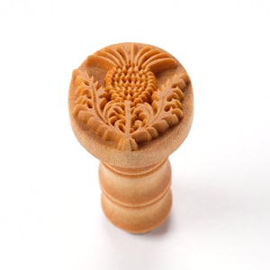 May include: A wooden stamp with a detailed carving of a thistle flower. The stamp is mounted on a wooden handle.