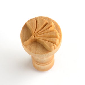 May include: A wooden stamp with a carved ginkgo leaf design. The stamp is light brown and has a round top with a cylindrical base.