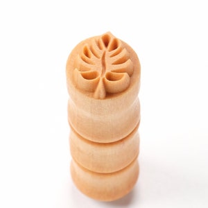May include: A wooden stamp with a leaf design. The stamp is made of light brown wood and has a round top with a leaf carved into it. The stamp is about 2 inches tall.