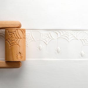 Könnte beinhalten: Ein hölzerner Nudelholz mit einem geschnitzten Spinnennetz- und Spinnendesign. Das Nudelholz ist hellbraun und weist ein sich wiederholendes Muster aus Spinnennetzen und Spinnen auf. Ideal für Backen und Basteln zum Thema Halloween.