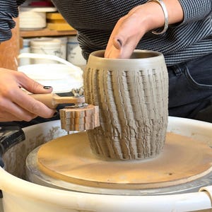 Puede incluir: Un recipiente cilíndrico de arcilla gris centrado en un torno de alfarero, con un patrón exterior texturizado creado por una herramienta de rodillo de madera con bordes dentados. La herramienta se presiona contra la arcilla para un diseño.