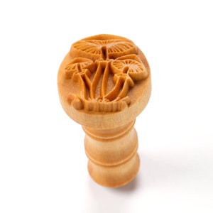 May include: A wooden cookie stamp with a design of three mushrooms. The stamp is brown and has a textured surface.