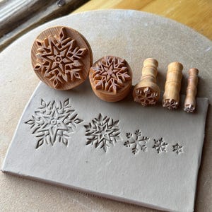 May include: A set of five light brown wooden stamps with snowflake patterns of varying sizes. The stamps are arranged next to a rectangular slab of light gray clay that features five corresponding snowflake impressions of different sizes.
