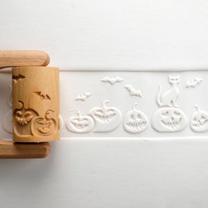 May include: A wooden rolling pin with carved Halloween-themed designs. The pin features pumpkins and bats. A white surface displays embossed pumpkins, bats, and a cat. This item is perfect for baking Halloween treats.