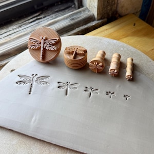 May include: A set of five round wooden stamps with dragonfly carvings of varying sizes, arranged in a row. Each stamp features a detailed insect design on the base, which is pressed into a flat, light gray slab of clay to create impressions.
