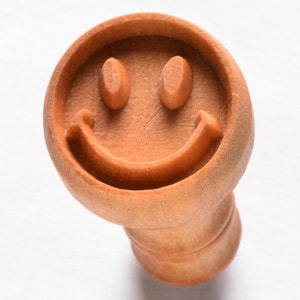 May include: A wooden smiley face cookie cutter. The cutter is brown and has a simple design.