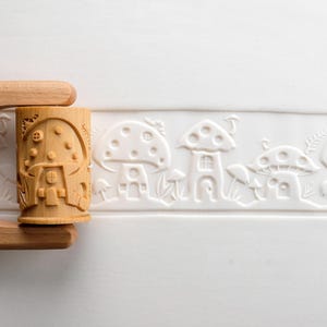 May include: A wooden rolling pin with a carved mushroom house design. The pin is light brown and features a repeating pattern of mushroom houses and other whimsical elements. The design is pressed into a white surface.