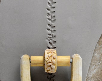 May include: A wooden rolling pin with a floral design is used to create an embossed pattern on a piece of clay.