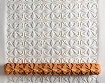 Op de afbeelding: Een houten deegroller met een gegraveerd geometrisch patroon. De deegroller ligt op een witte achtergrond met een vergelijkbaar geometrisch patroon.