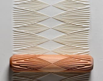 Op de afbeelding: Een houten deegroller met een gesneden ontwerp van overlappende waaiervormige vormen. De deegroller wordt gebruikt om decoratieve patronen in klei of deeg te maken.