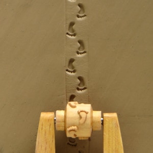 May include: A wooden rolling pin with a handle on each side. The rolling pin has a design of footprints on it. The rolling pin is being used to make footprints in clay.