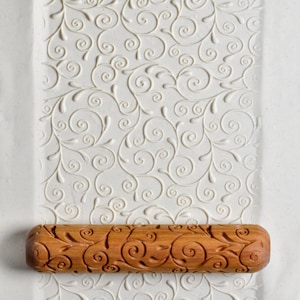 May include: A wooden rolling pin with a carved design of swirling vines and leaves. The rolling pin is on a white surface with a similar embossed design.