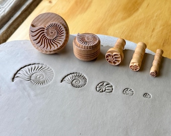 May include: A set of five light brown wooden stamps, each featuring a carved ammonite fossil design. The stamps vary in size from large to small, and each has created a corresponding spiral shell impression in the flat gray clay surface below.