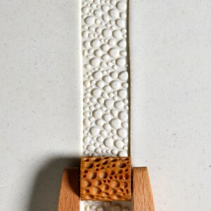 May include: A close-up of a wooden rolling pin with a honeycomb pattern. The rolling pin is on a white surface with a textured pattern of small circles.