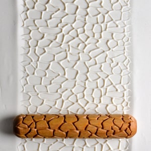 May include: A brown wooden rolling pin with a cracked-stone pattern, placed on a white surface with a matching embossed design. The rolling pin is designed for creating textured patterns on dough or clay. The rolling pin measures approximately 20 cm in length.