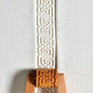 May include: A wooden roller stamp with a Celtic knot design. The roller is being used to create a pattern on a white surface.