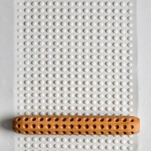 May include: A wooden rolling pin with evenly spaced holes rests on a white textured surface with a grid of small, raised dots. The rolling pin is a light brown colour and has a cylindrical shape.