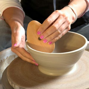 Op de afbeelding: Een lichtbeige kleikom gecentreerd op een ronde draaischijf. Een glad, ovaalvormig houten vormgereedschap wordt tegen de binnenwand van de kom gedrukt om de klei vorm te geven. De kom heeft een uitlopende rand en een taps toelopende voet.