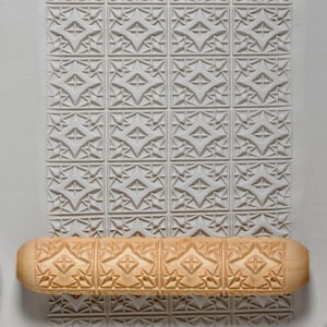 May include: A light-colored wooden rolling pin with a carved geometric pattern, next to a matching embossed pattern on a light gray surface. The rolling pin is cylindrical with rounded ends, and the pattern is repeated across its surface.