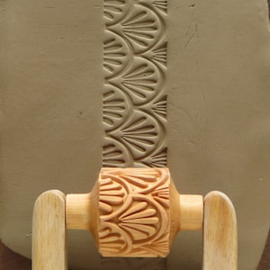 May include: A wooden rolling pin with a decorative pattern of overlapping fan shapes. The rolling pin is being used to create a textured design on a piece of clay.