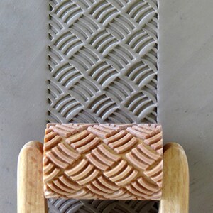 May include: A wooden rolling pin with a brown rubber roller featuring a repeating pattern of overlapping arches. The roller is used to create a textured design in clay.