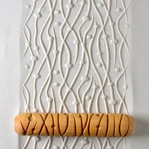 May include: A wooden rolling pin with a carved design. The rolling pin is on top of a white surface with a raised pattern of wavy lines and dots.