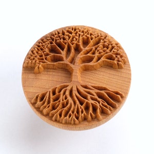 May include: A wooden round stamp with a detailed carving of a tree of life design. The tree has a thick trunk and branches with intricate details, and the roots are spread out in a circular pattern.