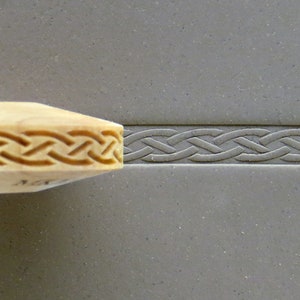May include: A wooden roller with a carved Celtic knot design. The roller is used to create a decorative border on clay or other materials.