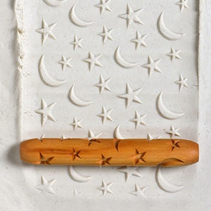 May include: A wooden rolling pin with a star and crescent moon design. The rolling pin is on a white surface with a pattern of stars and crescent moons.