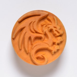 May include: A round, orange-colored object with a detailed relief carving of a dragon. The dragon is depicted with wings and a curved body, set against a plain white background. The carving has a textured appearance.
