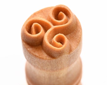 May include: A wooden stamp with a carved design of three interlocking spirals. The stamp is light brown and has a smooth, polished finish.