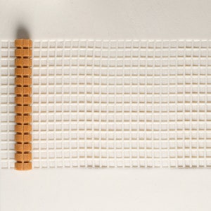 May include: A close-up of a white plastic mesh canvas with a brown wooden dowel rod on the left side. The mesh is made up of small squares.