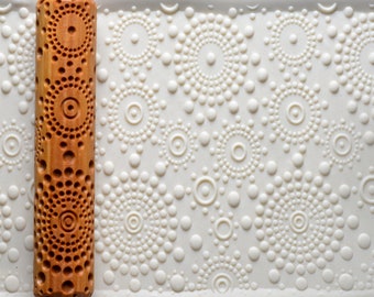 May include: A wooden rolling pin with a circular pattern of dots and rings. The rolling pin is on a white surface with a similar pattern.